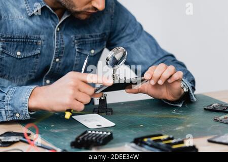 Beschnittene Ansicht des Reparaturwerkers, der durch die Lupe auf das zerlegte Teil schaut Des Mobiltelefons am Arbeitsplatz auf verschwommenem Vordergrund Stockfoto