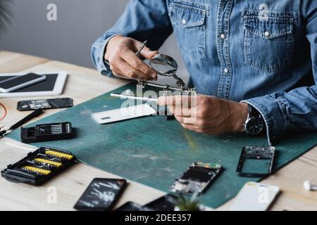 Beschnittene Ansicht des Reparaturwerkers, der zerlegte Teile des Mobiltelefons in der Nähe hält Lupe am Arbeitsplatz auf verschwommenem Vordergrund Stockfoto