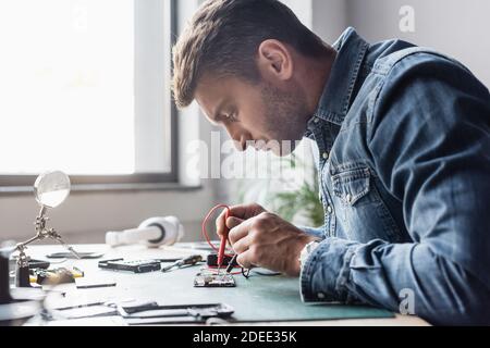 Seitenansicht des fokussierten Reparaturwerkers, der die Sensoren des Multimeters an hält Zerlegter Teil des Mobiltelefons auf verschwommenem Hintergrund Stockfoto