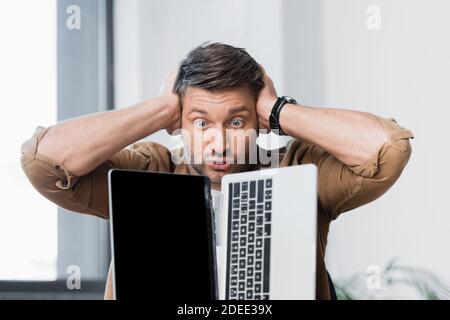 Mad Geschäftsmann mit Händen in der Nähe des Kopfes Blick auf beschädigte Laptop Auf unscharfem Hintergrund Stockfoto