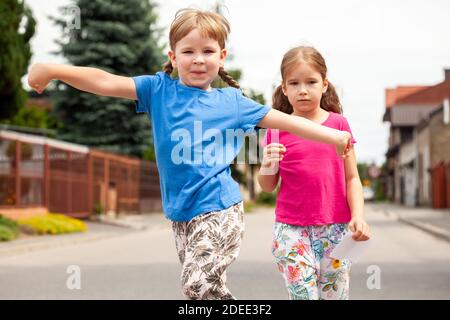 Zwei glückliche kleine Kinder, Schwestern, Geschwister gehen, Mädchen springen glücklich auf die Kamera mit weit ausgebreiteten Armen. Kinder Familienspaziergang, Stadtgebiet Stockfoto