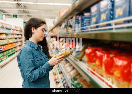 Einkaufen im Lebensmittelgeschäft. Eine junge Frau in einem Jeanshemd liest die Informationen auf einem Päckchen Pasta. Im Vordergrund verschwommene Regale mit Produ Stockfoto