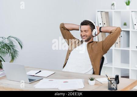 Fröhlicher Geschäftsmann mit Händen hinter dem Kopf Blick auf Laptop während Sitzen am Arbeitsplatz im Büro Stockfoto