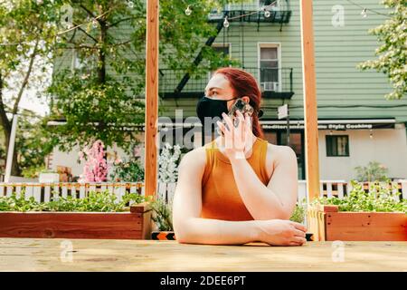 Junge Frau, die im Freien an einem Restauranttisch sitzt und Gesichtsmaske trägt. Stockfoto