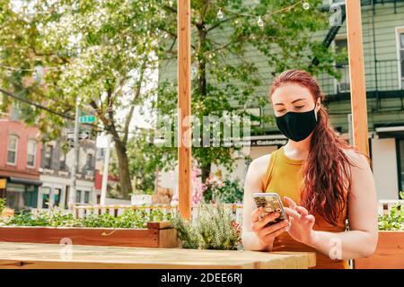 Junge Frau, die im Freien an einem Restauranttisch sitzt und Gesichtsmaske trägt. Stockfoto