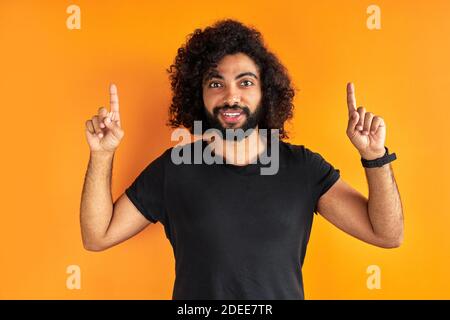 Optimistischer bärtiger junger Mann zeigt mit beiden Zeigefingern nach oben, optimistischer fröhlicher arabischer Kerl ist gut gelaunt, wirbt für neues Element, zeigt leer Stockfoto