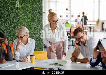 Wütende Chefin schimpft die Angestellten am Ende des Monats wegen eines schlechten Ergebnisses, schreit und schreit im Büro, diverse Arbeiter sitzen in Angst vor ihr Stockfoto