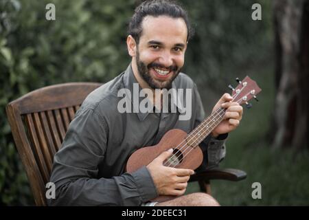 Ein glücklicher junger hispanischer bärtiger Mann, der mit einer Ukulele sitzt In einem Park Stockfoto