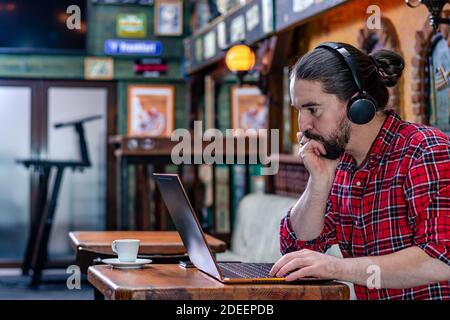 Portrait eines modernen jungen bärtigen Freiberuflers oder Bloggers, der in einer Bierstube sitzt, an einem Laptop arbeitet.Remote Work Konzept. Speicherplatz kopieren. Stockfoto