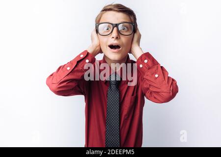 Portrait von positiven und emotionalen Schuljunge, posiert und bedeckt Ohren, weißer Hintergrund, Brille, rotes Hemd, Business-Thema Stockfoto