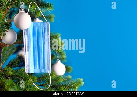 Medizinische Gesichtsmaske, die vom Weihnachtsbaum auf blauem Hintergrund mit leerem Kopierraum hängt. Konzept für Weihnachtsfeier in Zeiten des Corona Virus Stockfoto