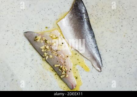 Die beiden Seiten einer Brasse in zwei Hälften geschnitten in Filets mit Petersilie, Knoblauch und Öl auf einem weißen Küche Schneidebrett mit schwarzen Punkten. Kochen Sie einfach. Stockfoto