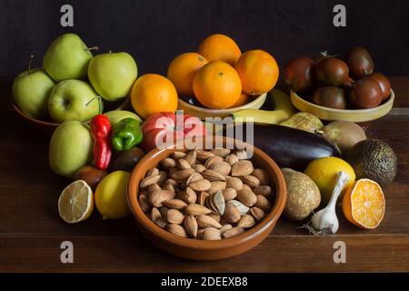 Stillleben Hintergrund mit ganzen Mandeln, Knoblauch, Banane, Orangen, Paprika, Avocado, Tomaten, Zitrone, Birnen und Äpfeln auf einem Tisch und Tonschüsseln. Stockfoto