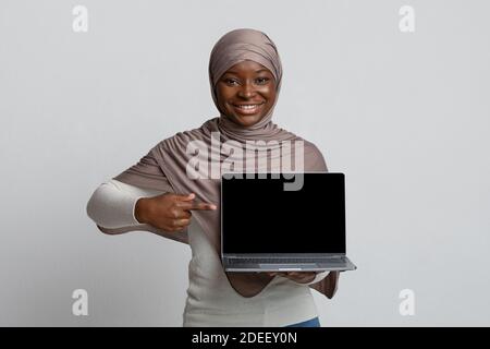 Afrikanische Dame In Hijab Laptop Halten, Zeigt Auf Schwarzen Bildschirm, Mockup Bild Stockfoto