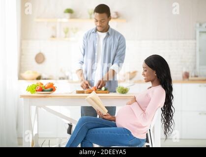 Afroamerikanische schwangere Frau liest Buch, während ihr Mann kocht Gemüsesalat in der Küche Stockfoto