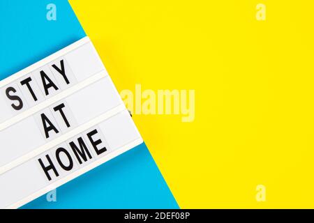 Leuchtkasten mit Text ZU HAUSE bleiben auf gelbem und hellblauem Hintergrund. Draufsicht Stockfoto