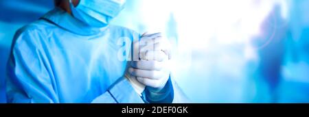 Frontline müde junge Ärztin setzen auf sterile Handschuhe. Tragen schützende medizinische Gesichtsmaske und blaue Uniform. Nahaufnahme der Hände. Breites Banner mit Kopierraum - Gesundheit, Medizin und Pandemie. Stockfoto