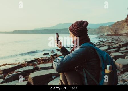 Ein junger Mann in warmen Kleidern sitzt auf dem Stein und fotografiert den Sonnenuntergang mit seiner Handykamera. Ruhen Sie sich bei einem Spaziergang an der felsigen Küste aus Stockfoto