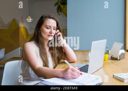 Junge Geschäftsfrau Notizen im Notebook machen, während am Telefon sprechen und arbeiten am Laptop in einem Café, Coworking. Remote-Arbeitsplatz. Freiberufler arbeitet Stockfoto