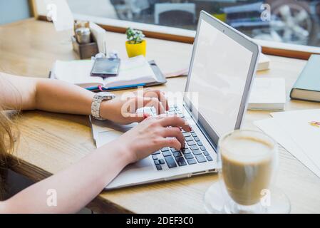 Nahaufnahme einer Frau, die an einem Laptop arbeitet und eine Tasse Kaffee trinkt, während sie in einem Café sitzt oder mit Blick auf die Straße zusammenarbeitet. Weicher, selektiver Fokus. Weiß Stockfoto