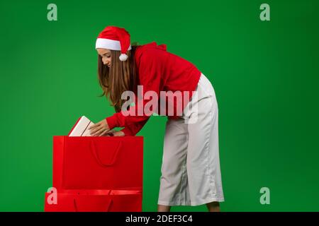 Ein junges Mädchen trägt einen weihnachtsmann Hut, zieht eine Geschenkbox aus einer großen Tasche. Grüner Hintergrund mit seitlichem Abstand. Stockfoto