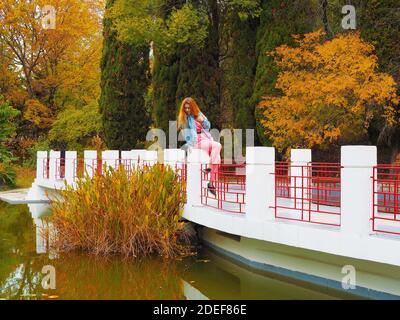 Rotschopf Mädchen sitzt auf dem Geländer der Brücke über Der Teich und schaut auf das Wasser im Park Stockfoto