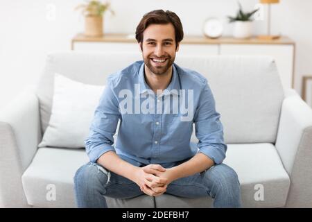 Glücklicher Mann, Der Auf Der Couch Sitzt Und Zu Hause Zur Kamera Lächelt Stockfoto