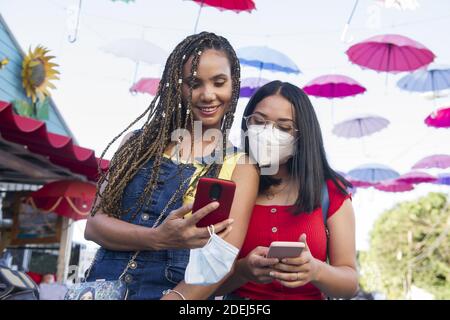 Halbkörperaufnahme von zwei attraktiven jungen hispanischen Mädchen mit Gesichtsmaske mit Smartphone in öffentlichen Straßen Platz in dominikanische republik mit bunten Dolden Stockfoto
