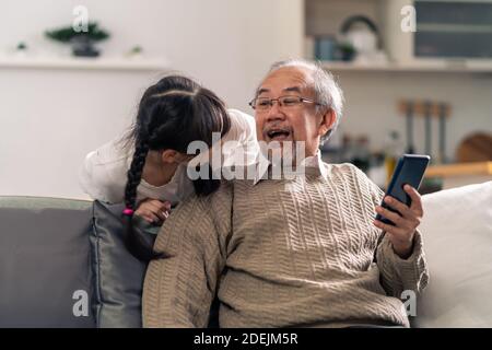 Glücklicher Ruhestand älterer Mann sitzt auf dem Sofa im Wohnzimmer mit Enkelin mit digitalen Tablet zusammen. Familie mit mehreren Generationen und Technologie Stockfoto