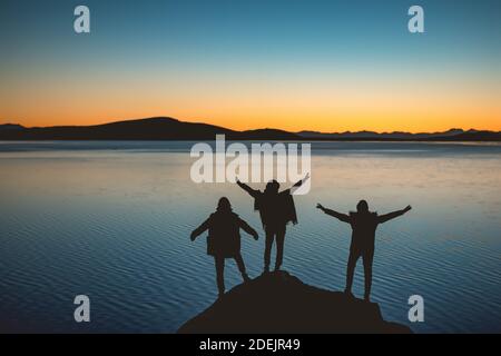 Silhouetten von drei glücklichen Mädchen mit erhobenen Armen steht dagegen Sonnenuntergang See und Berge Stockfoto