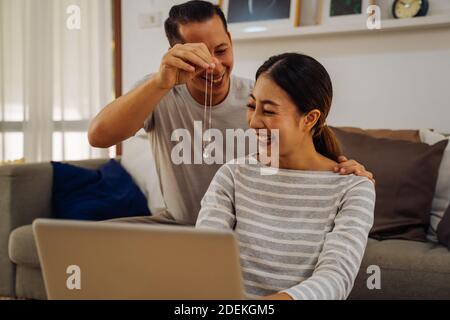 Junger Mann, der seiner Freundin eine Halskette als Geschenk gibt. Asiatische Frau immer überrascht von ihrem Freund mit Heiratsantrag während der Arbeit auf Laptop in modernen Hause Stockfoto