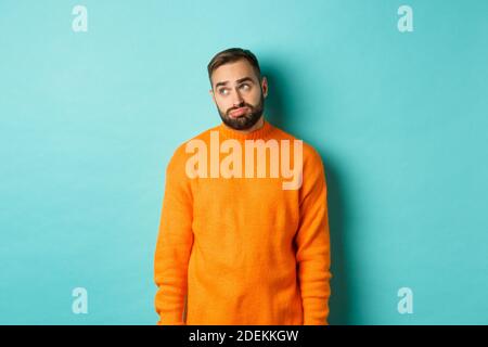 Trauriger und düsterer Mann schmollend, langweilen in der oberen linken Ecke, in orangefarbenem Pullover vor hellblauem Hintergrund stehend Stockfoto