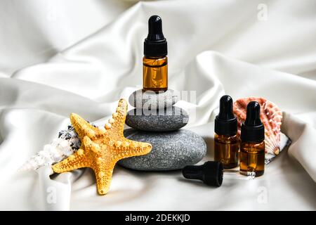 Kleine Flasche Serum auf Stapel von Basaltsteinen mit Muscheln und Seesterne auf Champagner-Satin-Stoff Hintergrund. Trendige Farben. Beauty Pipette Droppe Stockfoto