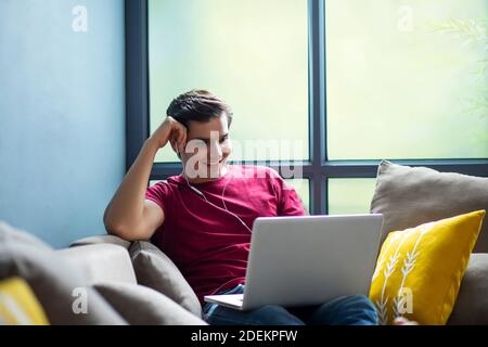 JUNGER MANN, DER SICH IM SITZEN EINEN FILM AUF SEINEM LAPTOP ANSIEHT AUF SEINER COUCH BEQUEM Stockfoto