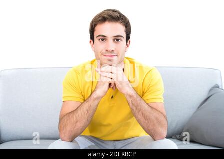 Junger gutaussehender lächelnder Kaukasischer Mann sitzt auf der Couch Wohnzimmer mit Blick auf die Kamera mit dem Kinn ruht auf Hände Stockfoto