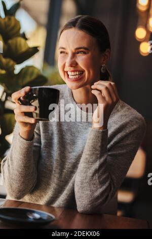 Frau hält Tasse Kaffee und lächelt. Fröhliche Frau, die in einem Café sitzt und Kaffee trinkt. Stockfoto
