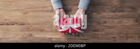 Frau in einem grauen Pullover geben ein weihnachts- oder Geburtstagsgeschenk in rotem Papier und weißem Band auf einem Holztisch gewickelt - Draufsicht, Panorama-Web-Banner w Stockfoto