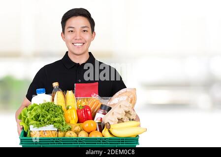 Lächelnder Lebensmittelladen asiatischer Liefermann trägt ein schwarzes Poloshirt Halten des Lebensmittelkorbs auf weißem Hintergrund mit Kopierplatz Stockfoto