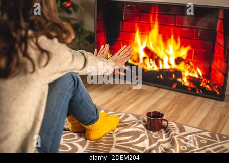 Frau in einem Pullover und gelben warmen Socken sitzt vor dem Kamin, der auf dem TV-Bildschirm dargestellt ist und wärmt ihre Hände. CR-Konzept Stockfoto
