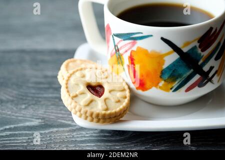 Ein jammy Dodger Keks mit seiner herzförmigen Mitte gefüllt mit Himbeermarmelade. Die Kekse, oder Kekse, auf einer Untertasse von einer Tasse mit einem heißen Getränk Stockfoto