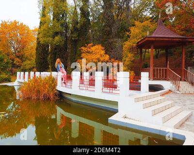 Rotschopf Mädchen sitzt auf einer Brücke über einem grünen Schlamm Teich in einem Park vor dem Hintergrund der Herbstbäume Und ein Pavillon Stockfoto