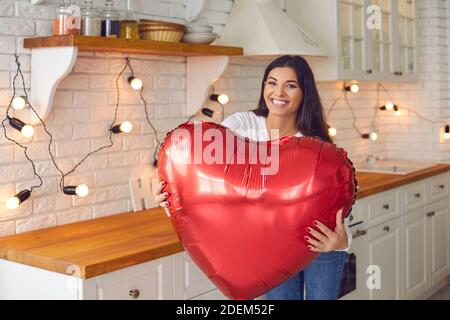 Glückliche junge Frau, die in der Küche steht, hält riesigen Valentine Ballon und lächelt Stockfoto