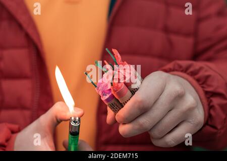 Mann in Rot aufgebockt Beleuchtung mehrere Feuerwerkskörper in seiner Hand mit Gasfeuerzeug. Guy immer bereit für das neue Jahr Spaß mit Feuerwerk oder pyrotechnische Produkte Stockfoto