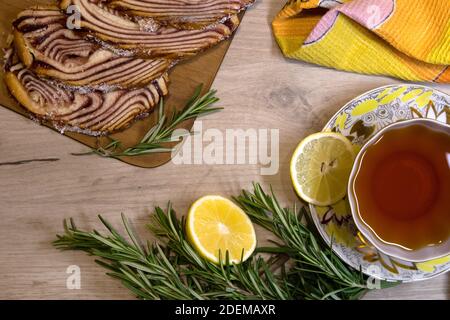 Das Frühstück wird auf einem Holztisch mit geschnittenem Gebäck, einer Tasse Tee, einer Zitronenhälfte und Rosmarinzweigen serviert Stockfoto