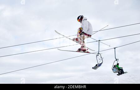 Seitenansicht des Mannes Skifahrer machen Sprung mit bewölktem Himmel und Skilifte im Hintergrund. Männliche Freerider auf Skiern springen in der Luft, während sie über schneebedeckte Pisten gleiten. Konzept des extremen Wintersports Stockfoto