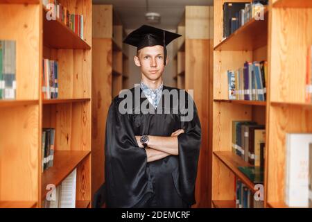 Erfolgreicher Absolvent Kerl, in akademischen Kleidern, posiert in der Bibliothek, kann für Werbung verwendet werden, Stockfoto