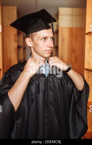 Erfolgreicher Absolvent Kerl, in akademischen Kleidern, posiert in der Bibliothek, kann für Werbung verwendet werden, Stockfoto