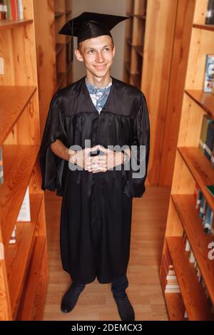 Erfolgreicher Absolvent Kerl, in akademischen Kleidern, posiert in der Bibliothek, kann für Werbung verwendet werden, Stockfoto