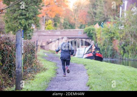 Rückansicht einer weiblichen Läuferin, die auf einem Fußweg entlang eines britischen Kanals läuft und sich einer Brücke nähert, an der ein Schmalboot in der Nähe festmacht. Stockfoto