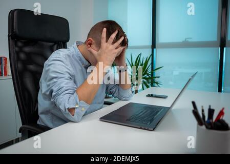Müde oder gestresste junge Geschäftsmann sitzt vor dem Computer im Büro. Unglücklicher Unternehmer am Arbeitsplatz. Ungesunder erschöpfter kaukasischer Mann havin Stockfoto